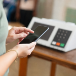 Pessoa usando celular para acessar serviços digitais da Justiça Eleitoral diante de uma urna eletrônica desfocada