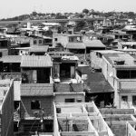 Vista aérea em preto e branco de um bairro urbano densamente povoado, com casas e telhados inacabados.